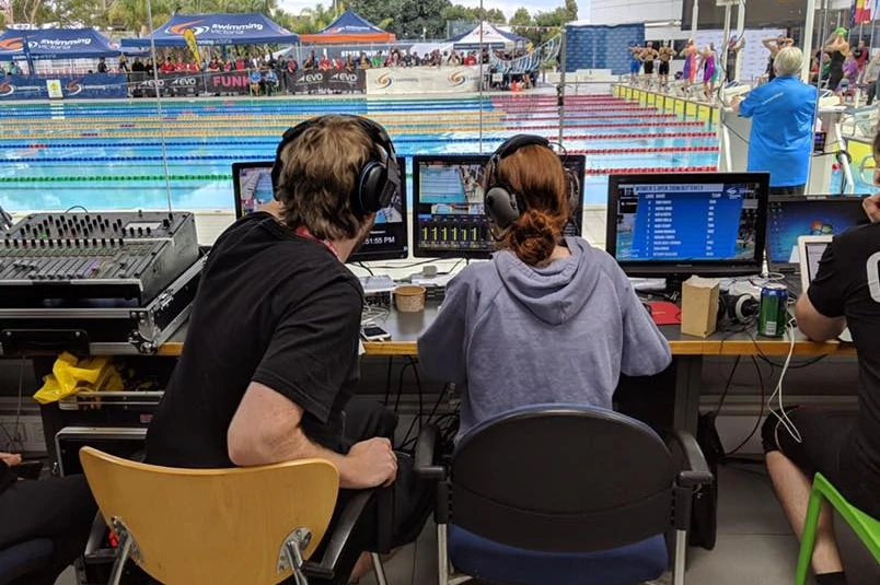 Three young people are looking at computers and, further on, a swimming pool where the competitors are getting ready to run.
