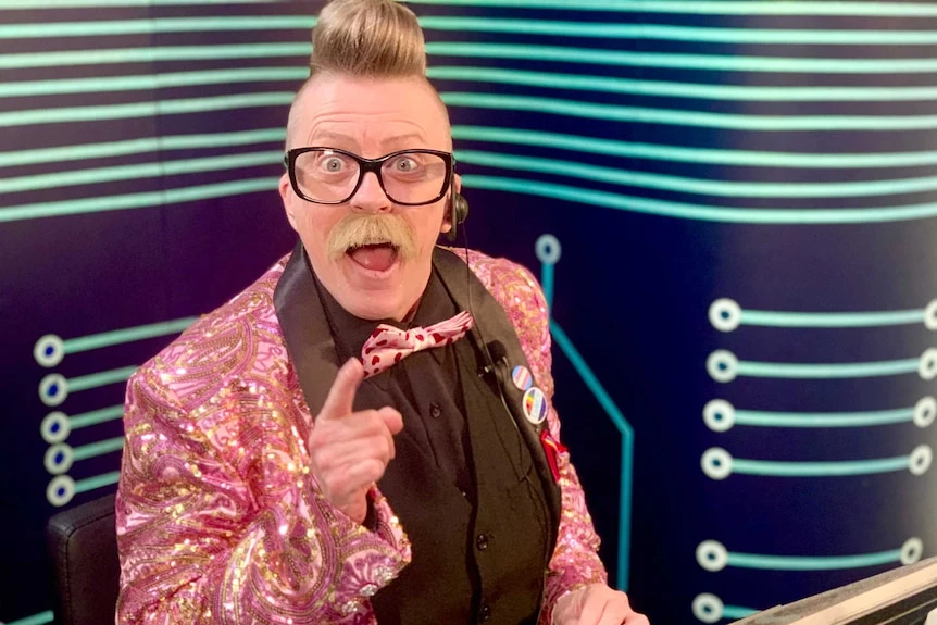 A man dressed in a sparkly pink jacket smiles at the camera with his finger pointed from a home TV studio